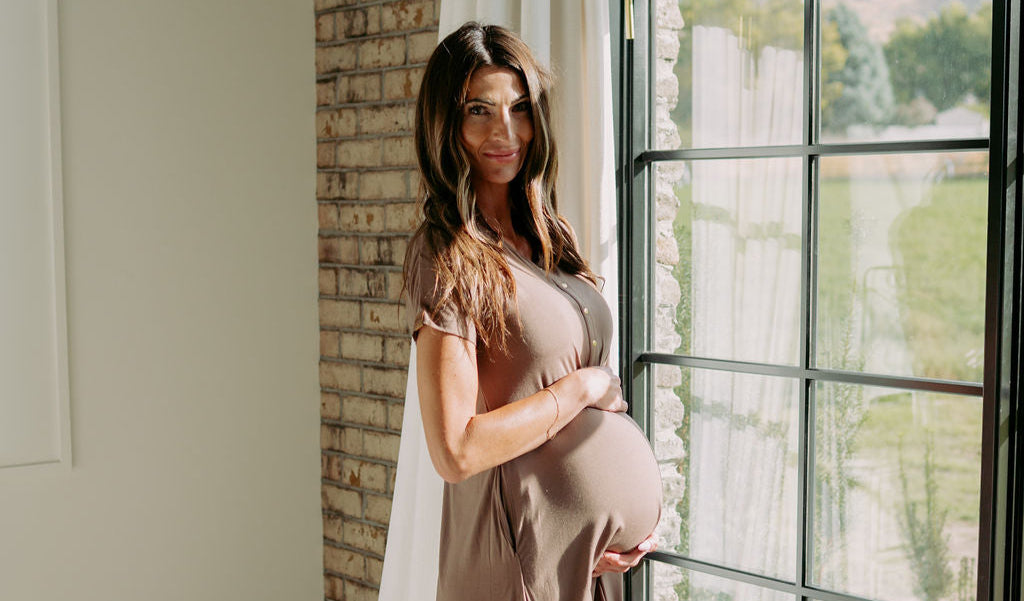 Pregnant woman in a brown dress standing by a large window with a view of greenery.